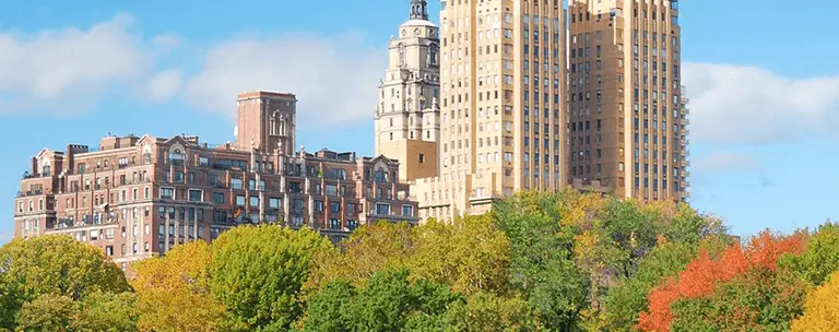 Panoramic view of historic buildings behind lush green and autumn-colored trees under a blue sky.