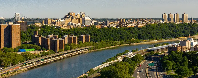 Aerial view of a river bordered by green trees and urban buildings under a clear sky.