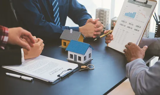 Three professionals in business attire at a table with house models, contract, keys, and financial report.
