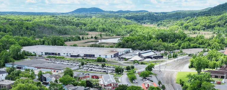 Aerial view of industrial buildings surrounded by greenery and distant hills under a partly cloudy sky.