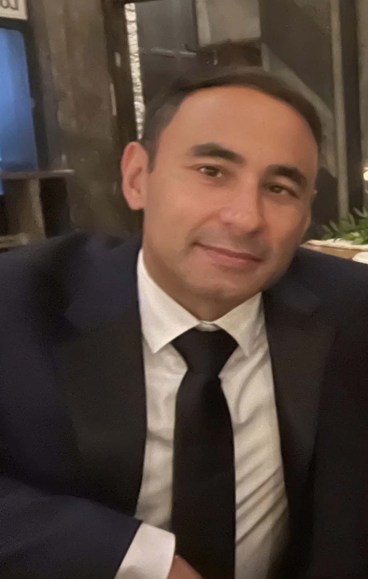 Portrait of a man in a dark suit, white shirt, and black tie, smiling softly indoors.