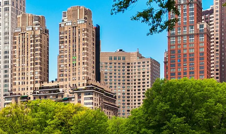 View of beige and red high-rise buildings behind lush green trees under a clear blue sky.