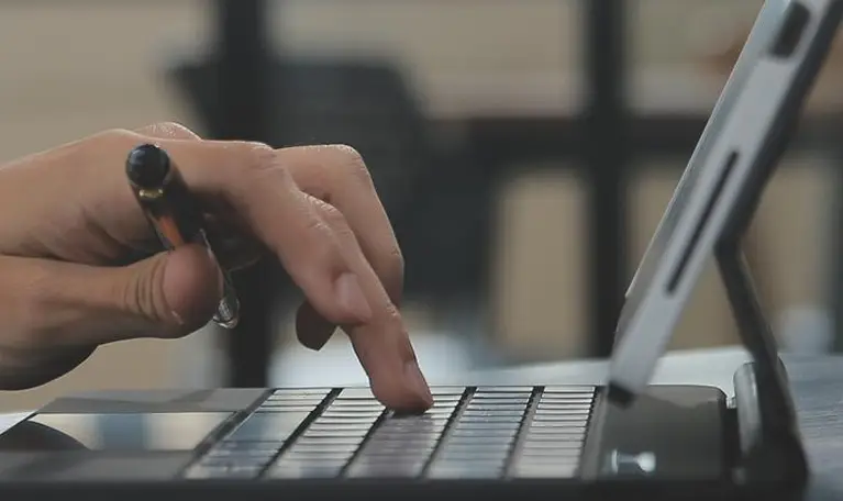 Close-up of a hand holding a pen and typing on a black laptop keyboard.