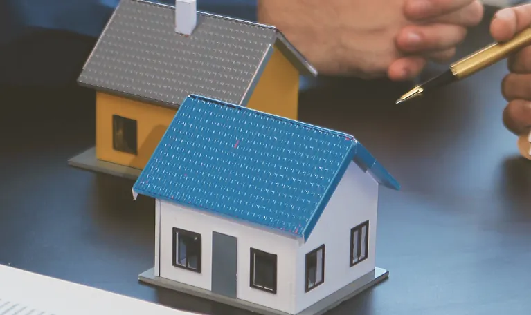 Two miniature house models, one white with a blue roof and one yellow with a gray roof, on a table with hands and a pen nearby.