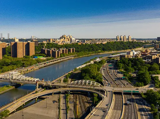 Aerial view of a river with multiple bridges, highways, green spaces, and urban buildings under a clear blue sky.