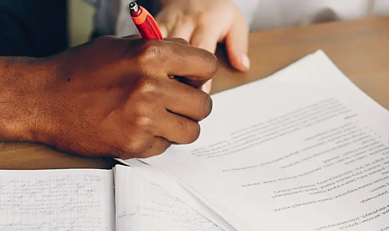 Close-up of a person’s hand holding a red pen, writing on printed documents with handwritten notes.