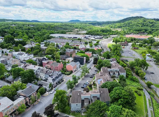 Aerial view of a suburban neighborhood with houses, trees, roads, and distant hills under a partly cloudy sky.
