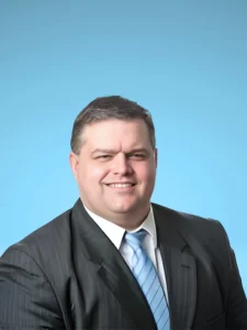 Portrait of a smiling man in a black suit and light blue tie against a blue background.