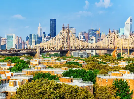 View of Queensboro Bridge with Manhattan skyline and residential buildings under clear blue sky.