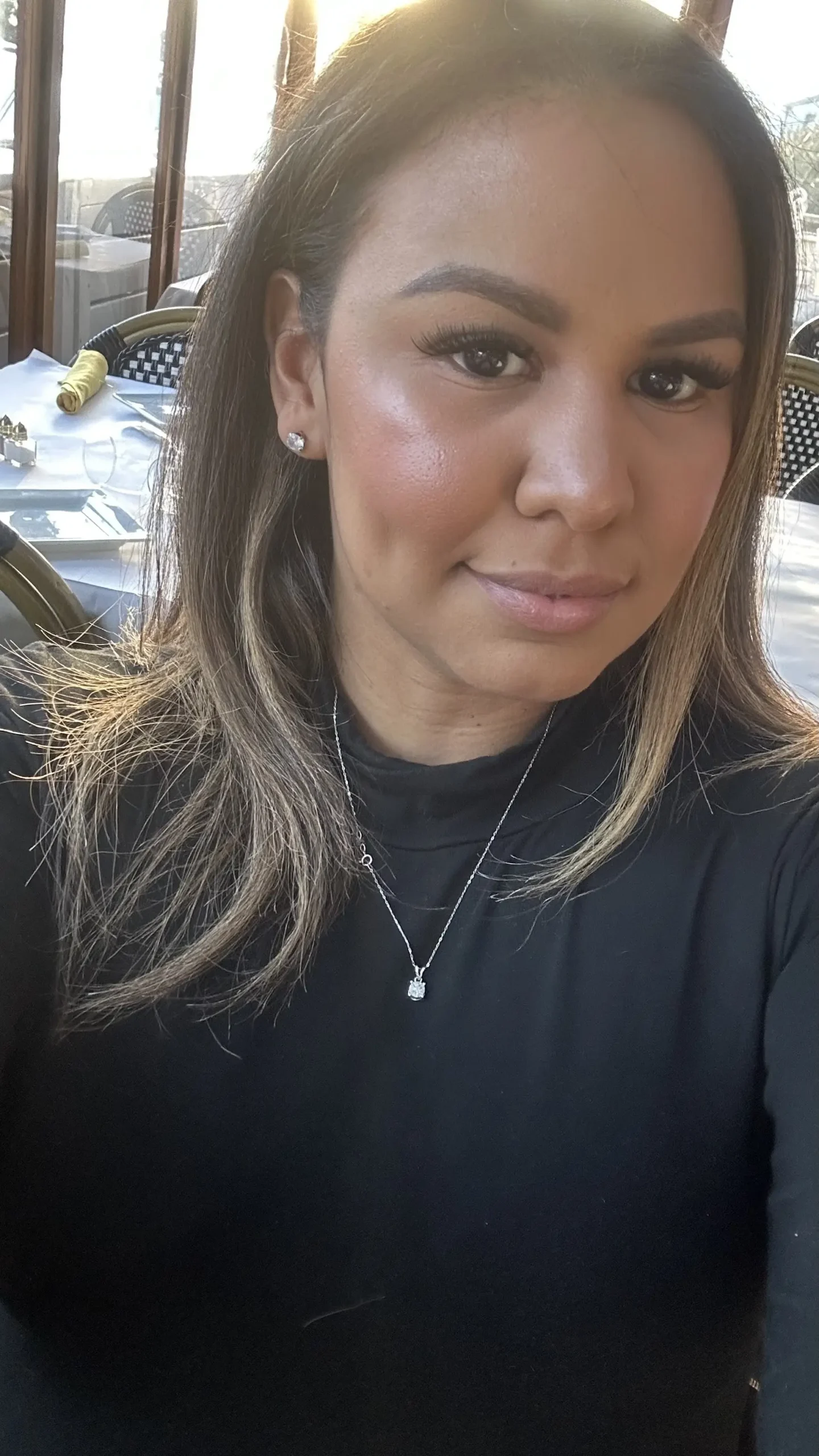 Close-up portrait of a woman with light brown hair, wearing a black top and diamond jewelry, seated at a restaurant table.