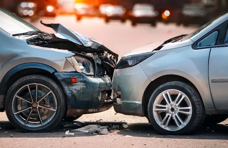 Two silver cars with front-end collision damage on a sunlit urban street.