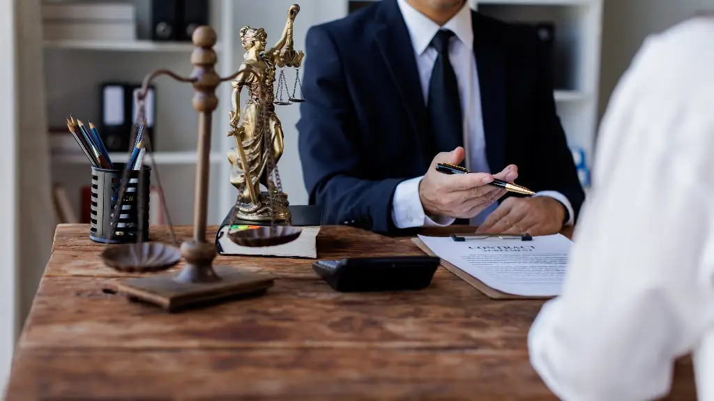 Two individuals in a legal consultation setting with a contract, scales of justice, and Lady Justice statue on a wooden desk.