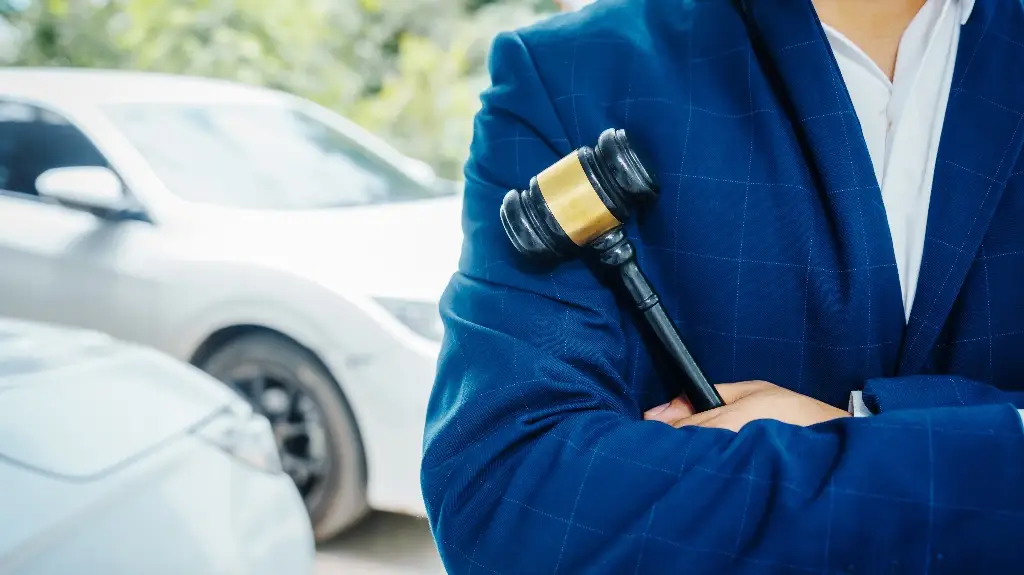 Person in blue suit holding black and gold gavel with two white cars in background.