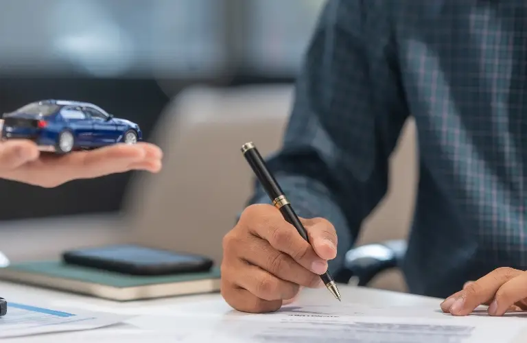 Person signing a document with a pen while another hand holds a blue toy car.