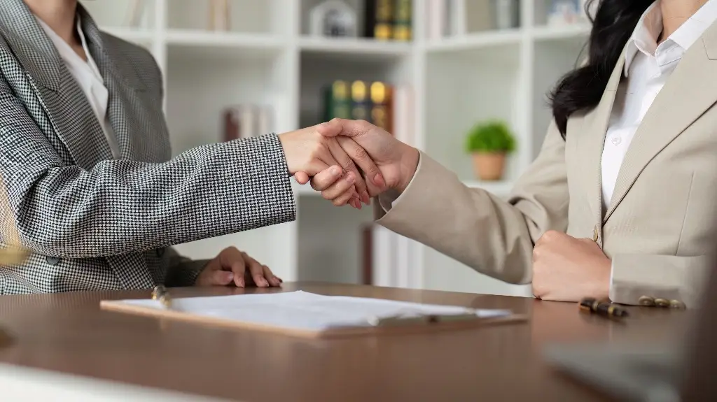 Two businesswomen shaking hands over a desk with documents in an office setting.