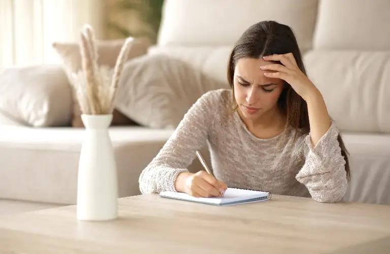 Young woman with long brown hair writing in a notebook at a beige wooden table, appearing focused and thoughtful, with a white vase containing dried plants and a light-colored sofa in the background.