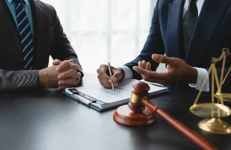 Two men in business suits discussing legal documents, with a gavel and scales of justice on the table.