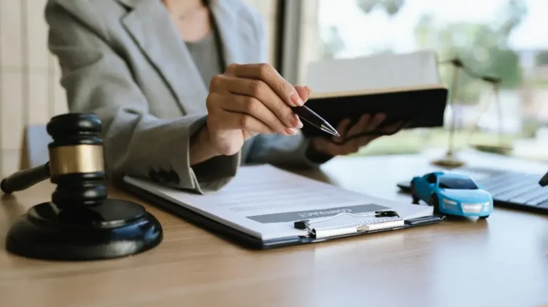 Person in gray suit holding pen and book over a contract on clipboard with gavel and toy car on desk.