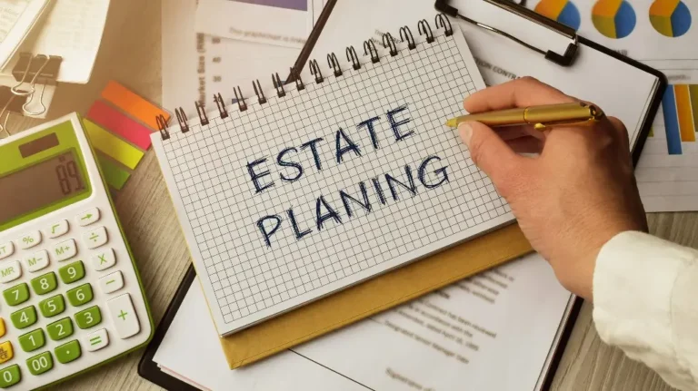 Hand writing "ESTATE PLANNING" on a spiral notebook with financial documents, calculator, and charts on a wooden desk.
