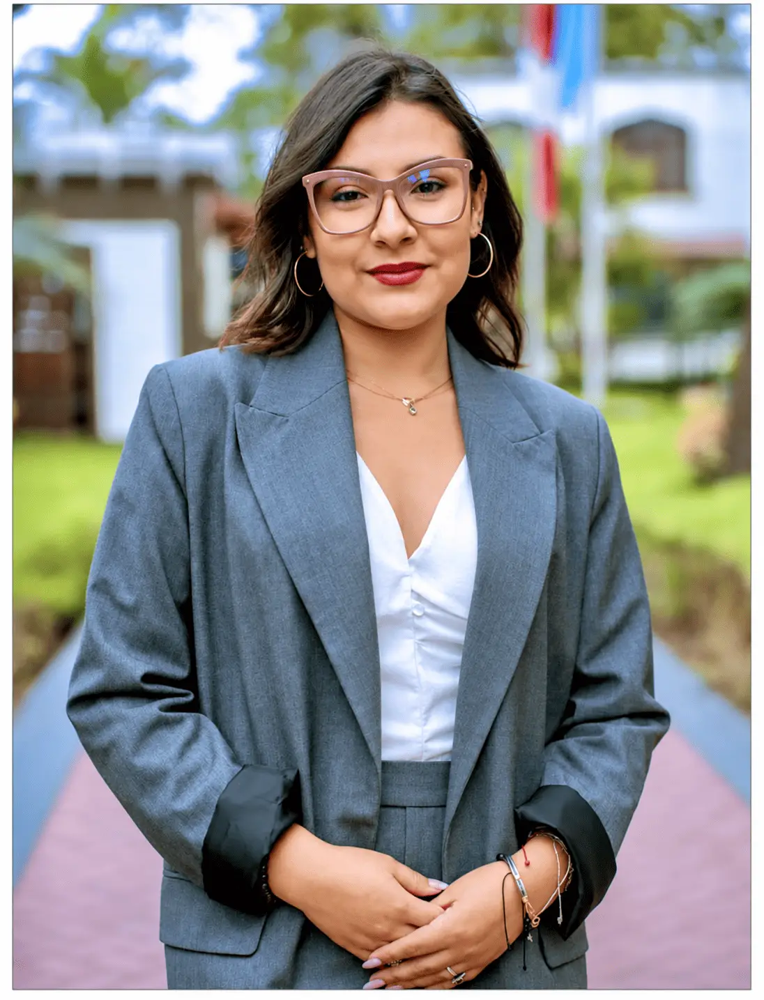 Professional woman in grey suit and glasses standing outdoors with hands clasped.